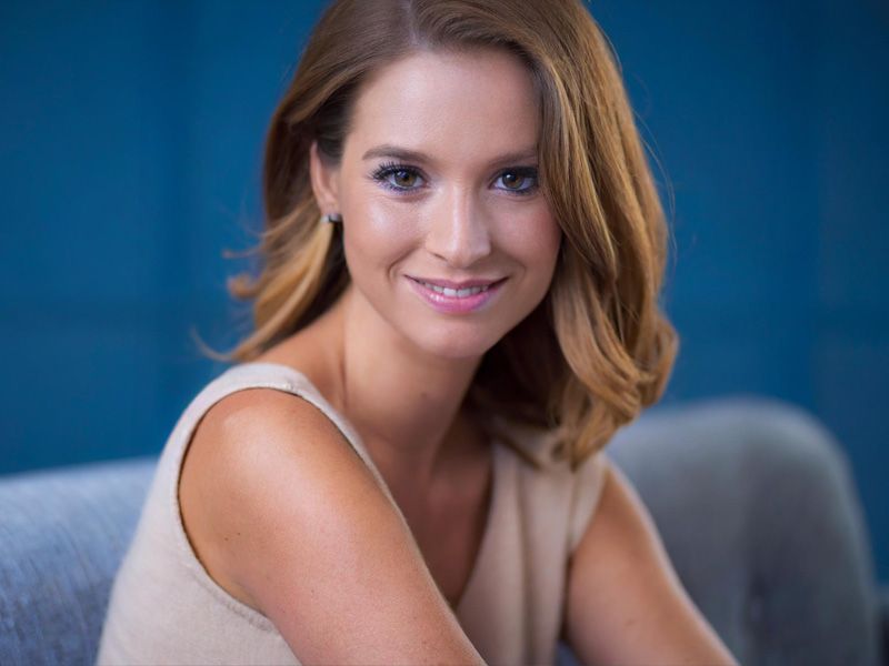 Woman smiling softly while seated, showing smooth skin and a natural, youthful complexion in a soft blue studio setting.