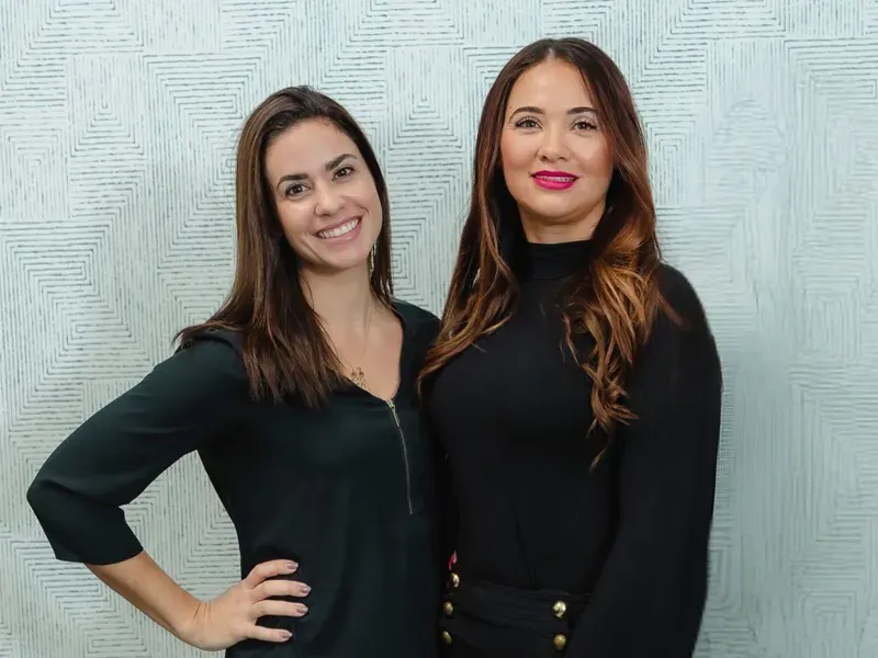 Sandra and Juliana standing together and smiling in front of a light patterned background, showing a friendly and professional appearance.